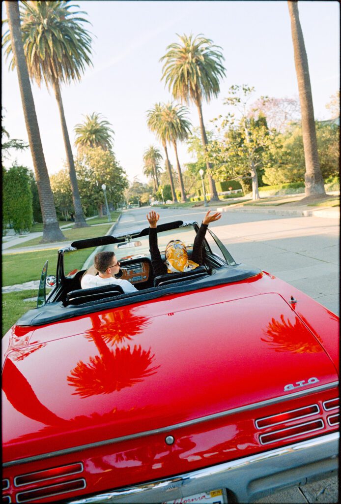Red convertible driving down palm tree lined street with woman's hands in the air